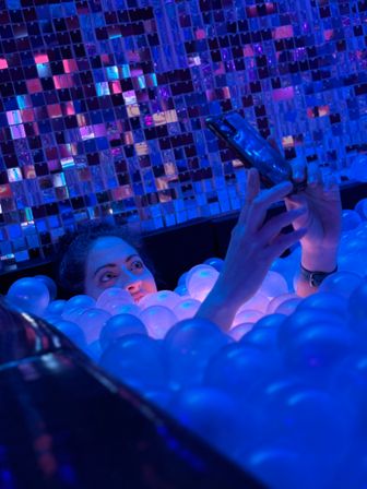 Person peeking from a neon blue and purple illuminated ball pit, taking a smartphone selfie in front of a mirrored mosaic wall at an indoor interactive art installation.