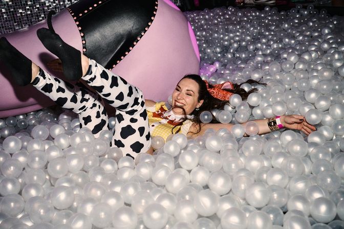 Woman laughing and lounging in a translucent white indoor ball pit at a party, wearing cow-print leggings, yellow top, black socks and a pink bandana with a large pink inflatable in the background.