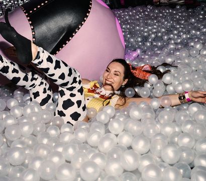 Woman laughing and lounging in a translucent white indoor ball pit at a party, wearing cow-print leggings, yellow top, black socks and a pink bandana with a large pink inflatable in the background.