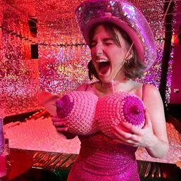 Person in a sparkly pink dress and mirrored cowboy hat laughing while holding knitted novelty breasts in a glittery pink disco room with a ball pit.
