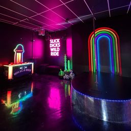 Neon-soaked nightclub interior with a pole performance stage, rainbow LED arch, glowing wall slogan, arcade game and glossy black floor.
