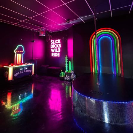 Neon-soaked nightclub interior with a pole performance stage, rainbow LED arch, glowing wall slogan, arcade game and glossy black floor.