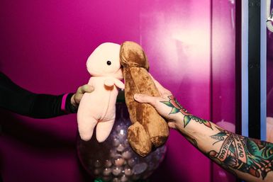 Two hands holding novelty plush toys — a small pink character and a brown novelty-shaped toy — touching in front of a bright magenta background, one arm tattooed.
