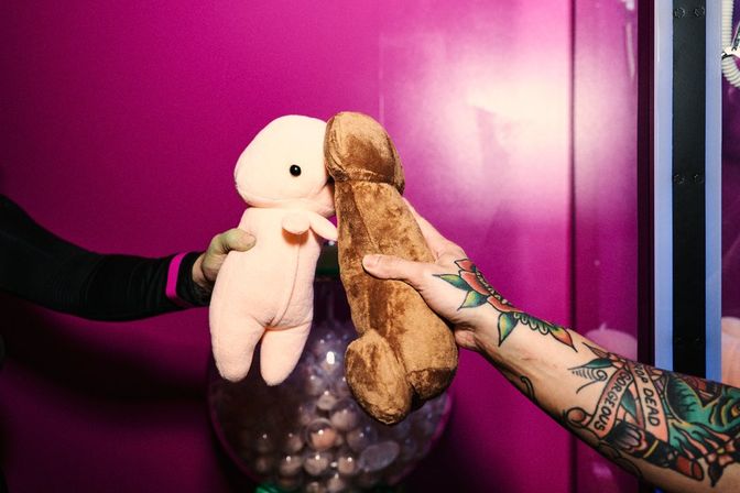Two hands holding novelty plush toys — a small pink character and a brown novelty-shaped toy — touching in front of a bright magenta background, one arm tattooed.