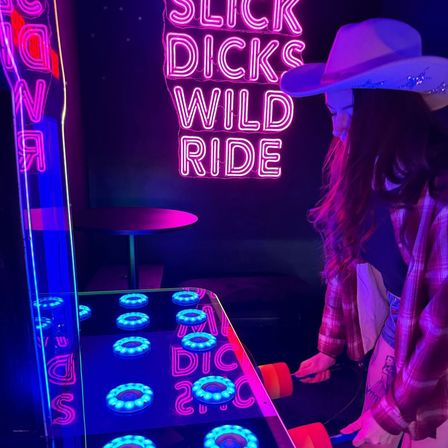 Person in a cowboy hat playing a glowing whac-a-mole style arcade game in a neon-lit bar arcade, pink-and-blue lights and a neon sign reading 'SLICK DICKS WILD RIDE'.