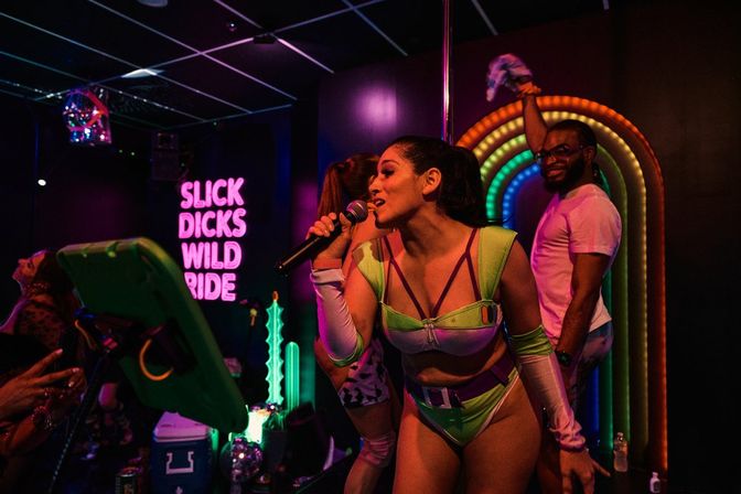 Neon-lit nightclub stage with a cabaret performer in a green futuristic costume singing into a microphone by a pole, rainbow arch lights and an animated crowd in the background.