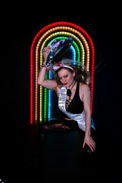 Playful party portrait of a woman in a black outfit and sparkly leggings, wearing a white celebration sash and veil, tipping a mirrored disco hat in front of a neon rainbow arch backdrop.