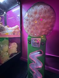 Neon-lit arcade gumball machine with a green token-operated base, clear globe packed with white plastic capsules and a spiral chute, next to a plush toy claw machine under pink-purple lighting.
