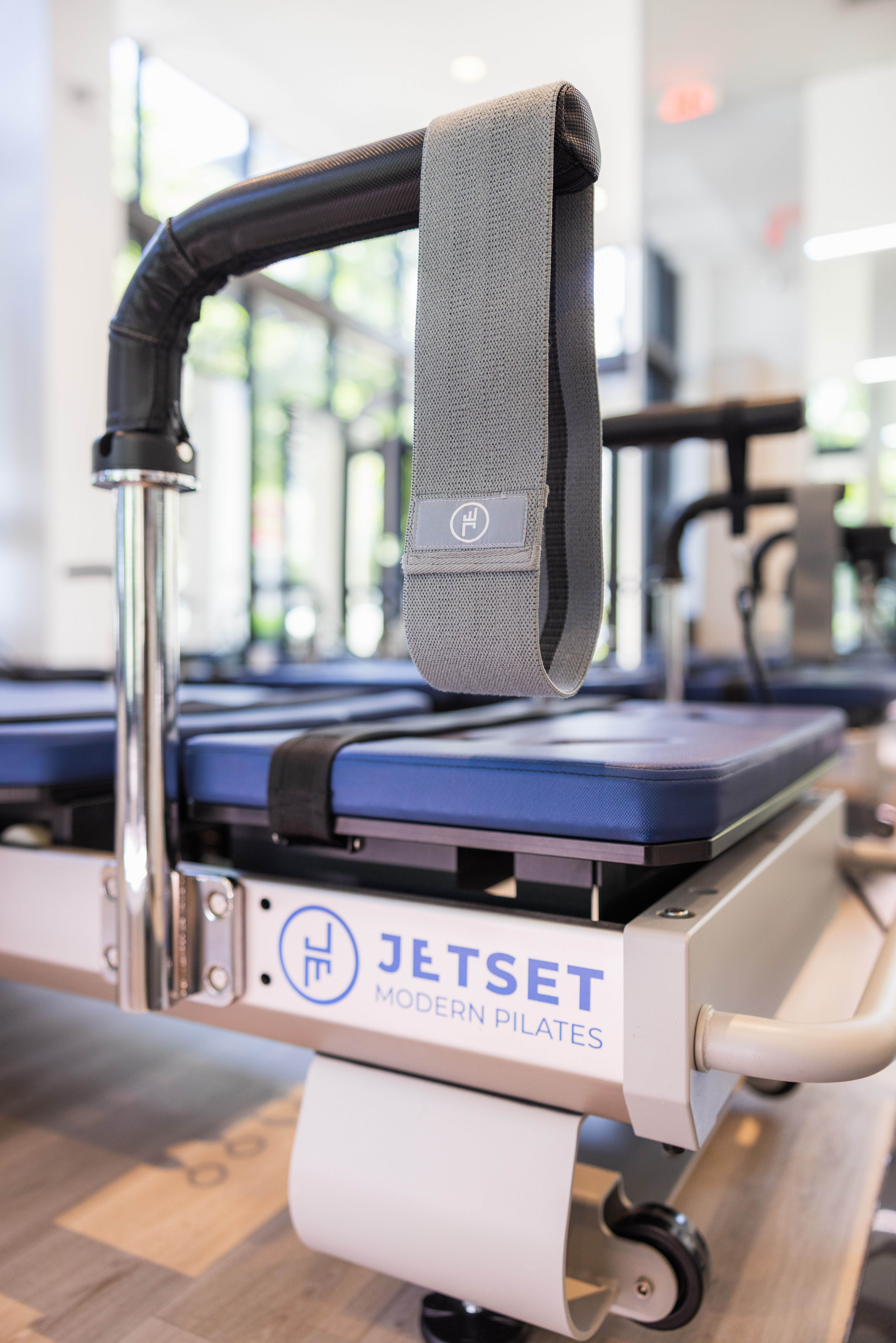 Close-up of a gray fabric strap hanging from a Pilates reformer machine above a blue padded carriage in a bright modern studio, fitness equipment detail