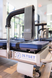 Close-up of a gray fabric strap hanging from a Pilates reformer machine above a blue padded carriage in a bright modern studio, fitness equipment detail