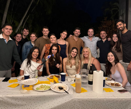 Smiling group of friends at a nighttime tropical backyard patio dinner, gathered around a long table with plates, yellow napkins, drinks and a paper towel roll — one person playfully using a small kitchen torch, casual outdoor party vibe.
