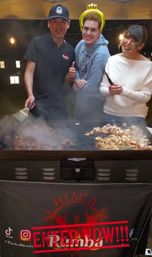 Three people smiling behind a steaming outdoor hibachi griddle at a nighttime food pop-up — cook with spatula, friend wearing an inflatable crown, and guest holding tongs while mushrooms and diced meat sizzle on the flat-top.
