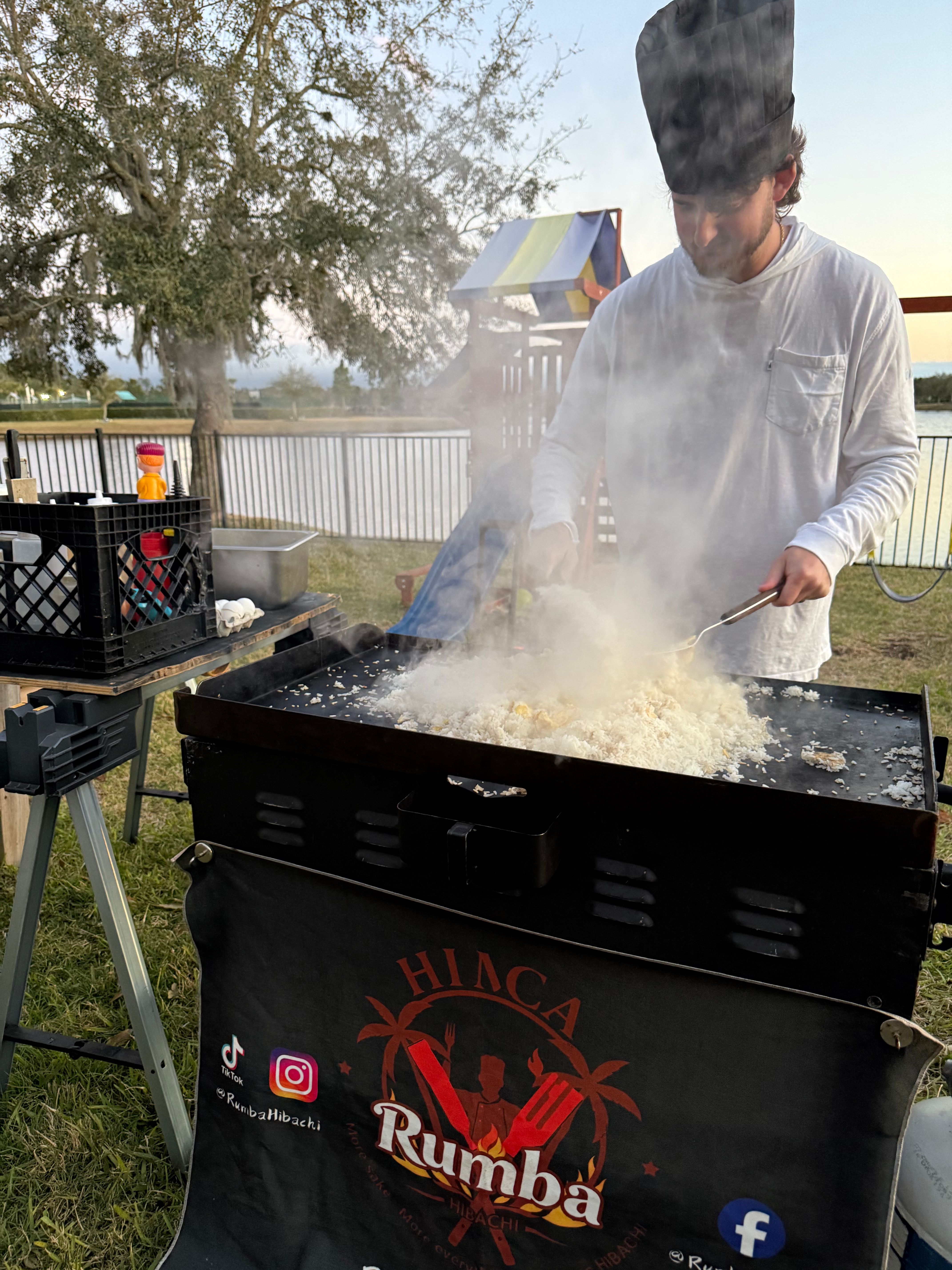Lakeside outdoor hibachi cook wearing a tall chef hat, stirring steaming fried rice on a flat-top griddle at sunset with a prep crate and playground in the background.