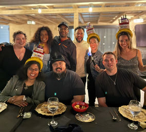 Group of nine friends smiling at a birthday dinner in a rustic indoor dining space, three wearing cake-shaped 'Happy Birthday' hats, gathered around a table with plates, wine glasses and desserts as one person holds a small celebratory torch.
