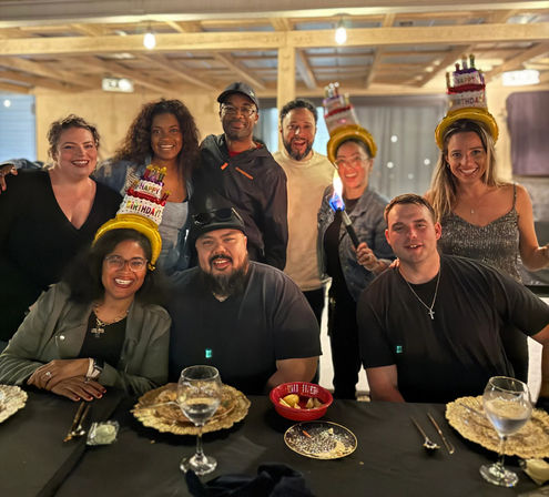 Group of nine friends smiling at a birthday dinner in a rustic indoor dining space, three wearing cake-shaped 'Happy Birthday' hats, gathered around a table with plates, wine glasses and desserts as one person holds a small celebratory torch.