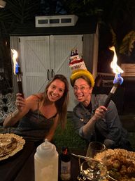 Two friends at a backyard birthday party at night, one wearing a cake-shaped birthday hat and both holding flaming torches, smiling over a table of food and drinks in a garden setting.