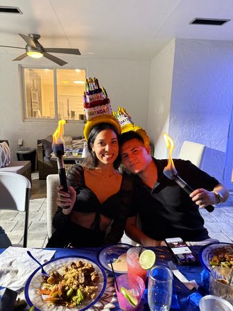 Smiling pair wearing inflatable "Happy Birthday" cake hats hold lit tiki torches at an evening backyard patio birthday dinner with plates of food and colorful drinks.