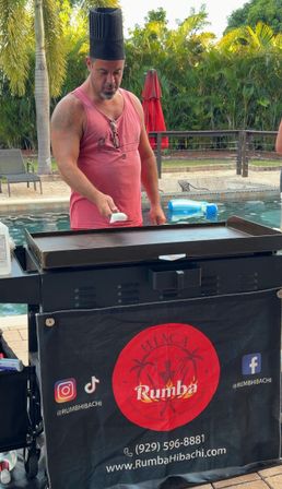 Poolside cook in a tall black chef hat and pink tank top working a flat-top griddle in a tropical backyard with palm trees, lounge chairs and a red umbrella.