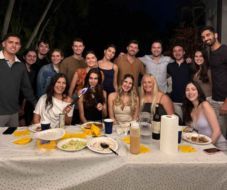Smiling group of friends at an outdoor nighttime dinner party on a patio, about 17 people gathered behind a long polka-dot table with plates, cups, drinks and condiments for a casual festive get-together.
