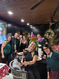 Smiling group of eight adults at a covered outdoor patio holiday party with wooden slat wall and wreath, plates of food on a counter, a woman in front holding two lit torches and another wearing an inflatable bunny-ear headband.