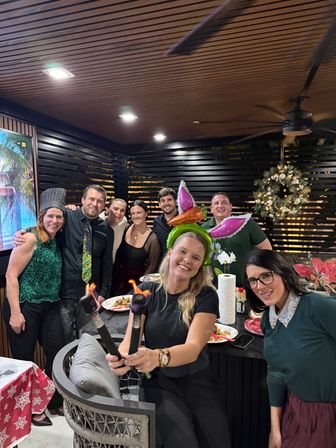 Smiling group of eight adults at a covered outdoor patio holiday party with wooden slat wall and wreath, plates of food on a counter, a woman in front holding two lit torches and another wearing an inflatable bunny-ear headband.