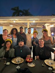 Nine friends celebrating a birthday on an outdoor patio at night under a wooden pergola with hanging lights, three people wearing novelty "Happy Birthday" cake hats, table set with plates, wine glasses and small bowls, tropical palm trees silhouetted in the dark sky.