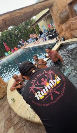 Tropical backyard pool party at a rock grotto: a man in a black shirt leans over the spa while friends in swim trunks relax in the pool, with colorful deck chairs and palm trees in the background.