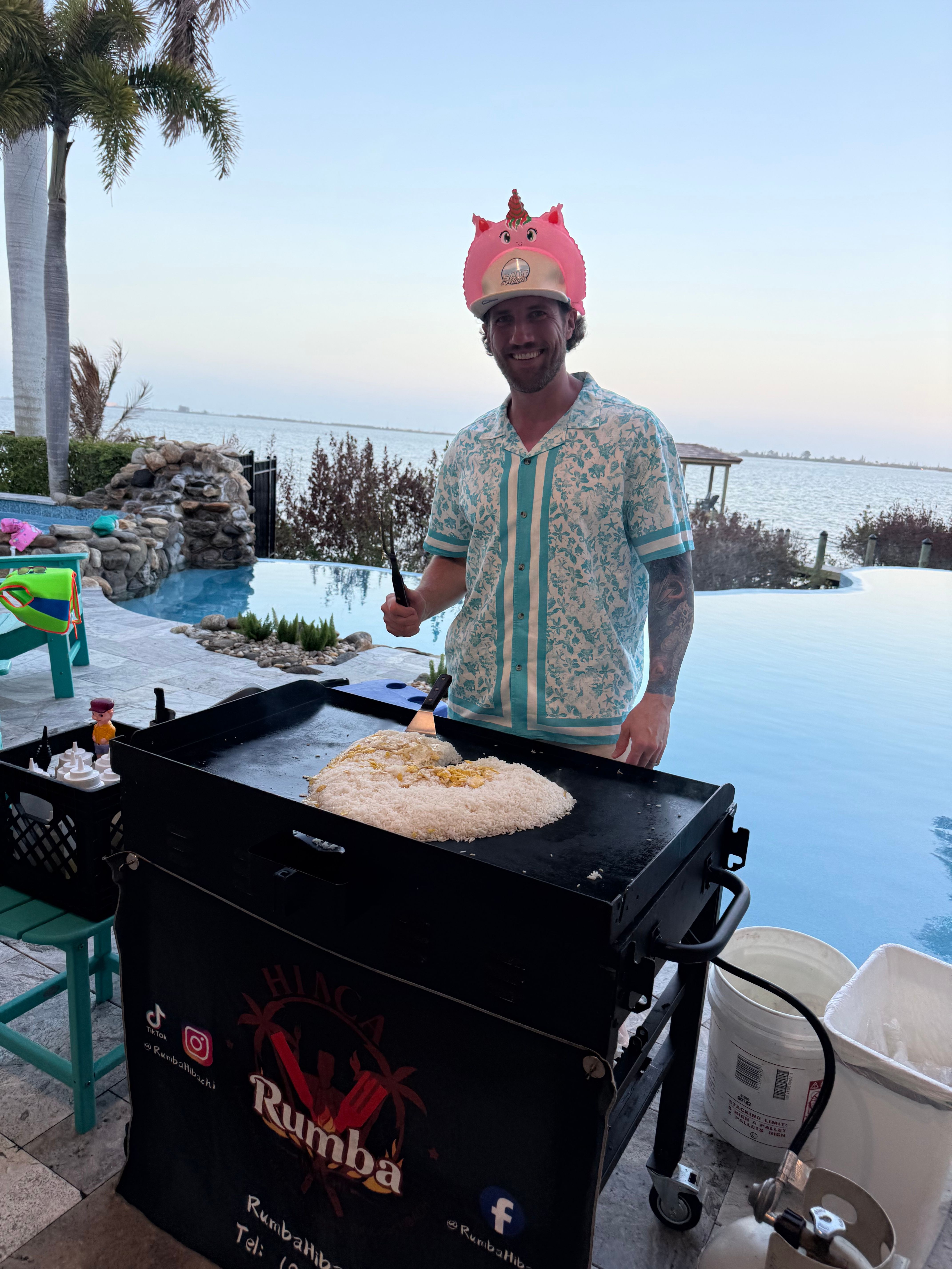 Person in a tropical shirt wearing a pink inflatable unicorn helmet cooking a large mound of fried rice on an outdoor griddle at a poolside waterfront patio with bay view