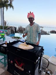 Person in a tropical shirt wearing a pink inflatable unicorn helmet cooking a large mound of fried rice on an outdoor griddle at a poolside waterfront patio with bay view