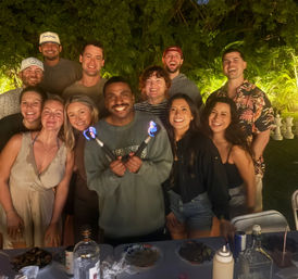 Smiling group of friends at a nighttime backyard party in a tropical garden, center person holding two small flaming torches above a table with drinks and plates.