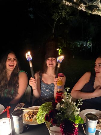 Three friends laughing at a nighttime backyard dinner party, center person holding two lit tiki torches, floral centerpiece and plates of food on the table with string lights wrapped in a tree overhead.