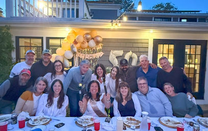 Smiling adults at an evening backyard birthday party on a suburban patio, gathered around a long dinner table with plates and red cups, gold-and-white balloons with silver '50' numbers and string lights overhead.