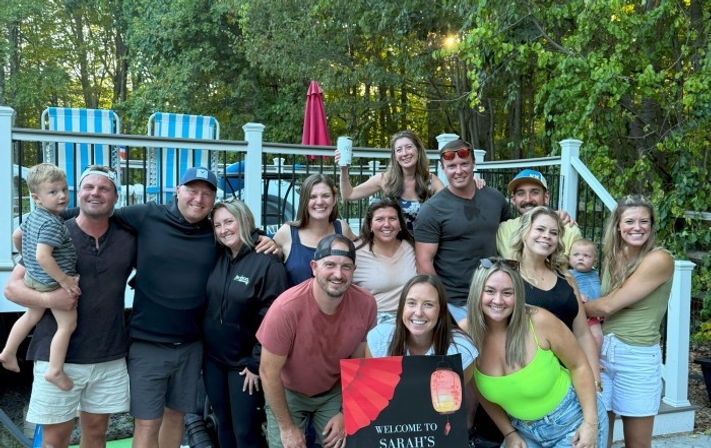 Smiling group of friends and family on a backyard pool deck at sunset, casual summer gathering with children, striped lounge chairs, umbrella and wooded yard in the background, holding a welcome sign.