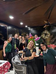 Smiling group at a festive holiday party on a covered outdoor patio — woman in front wearing a green bunny‑ear headband holding two small flaming torches, wreath and poinsettia decorations visible.
