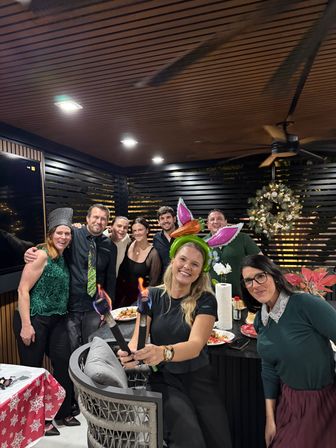 Smiling group at a festive holiday party on a covered outdoor patio — woman in front wearing a green bunny‑ear headband holding two small flaming torches, wreath and poinsettia decorations visible.