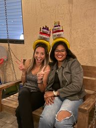 Two friends seated on a wooden patio swing bench wearing inflatable “Happy Birthday” cake hats, smiling and flashing peace signs during a birthday celebration.