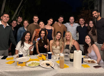 Smiling group of friends at a nighttime backyard dinner party under palm trees, gathered around a long table with plates, drinks, napkins and a handheld culinary torch.