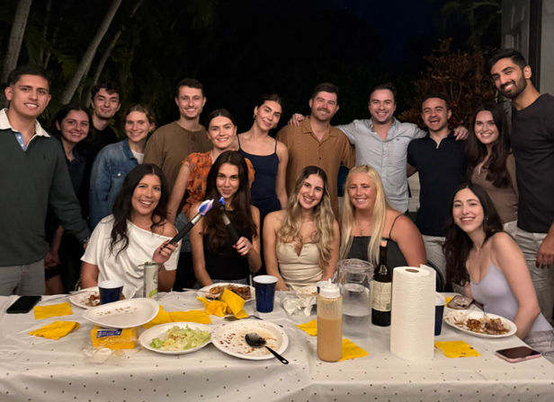 Smiling group of friends at a nighttime backyard dinner party under palm trees, gathered around a long table with plates, drinks, napkins and a handheld culinary torch.