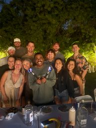 Smiling group of friends at an evening backyard garden party under lush tropical trees, gathered around a table with drinks and plates while the person in front holds two small flaming torches.