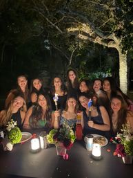 Nighttime outdoor garden dinner with a smiling group of friends gathered around a candlelit table, holding lit torches and surrounded by floral centerpieces and tree branches wrapped in string lights — warm, celebratory atmosphere.