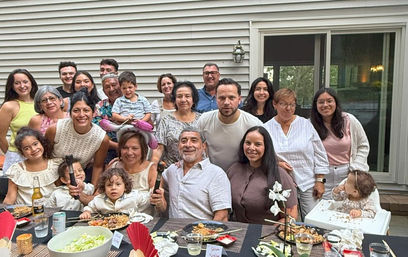 Cheerful multigenerational family of about 18 posing at an outdoor patio dinner in a suburban backyard, gathered around a table of food in front of a house with vinyl siding and sliding glass door.