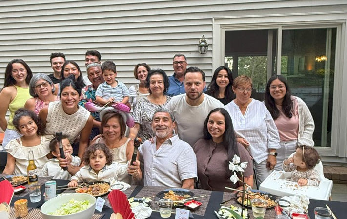Cheerful multigenerational family of about 18 posing at an outdoor patio dinner in a suburban backyard, gathered around a table of food in front of a house with vinyl siding and sliding glass door.