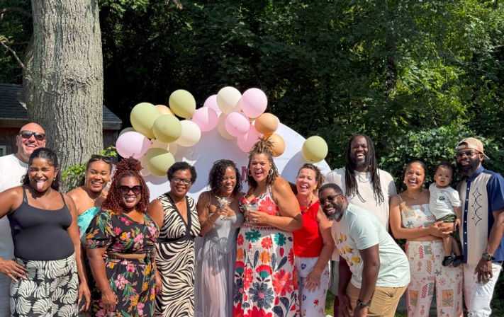 Smiling multi-generational group posing under a pastel balloon arch at a sunny backyard celebration, adults in colorful summer outfits and one toddler—festive garden party vibe.