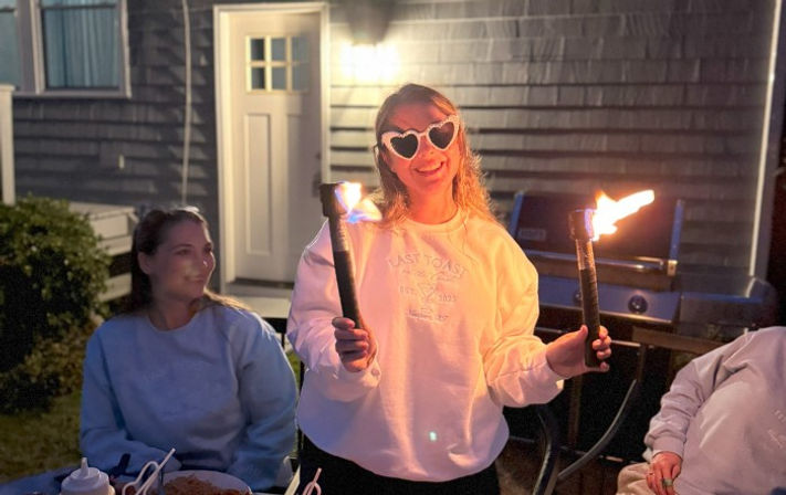 Smiling person wearing heart-shaped sunglasses holding two lit tiki torches at a backyard patio gathering by a grill and house door at night.