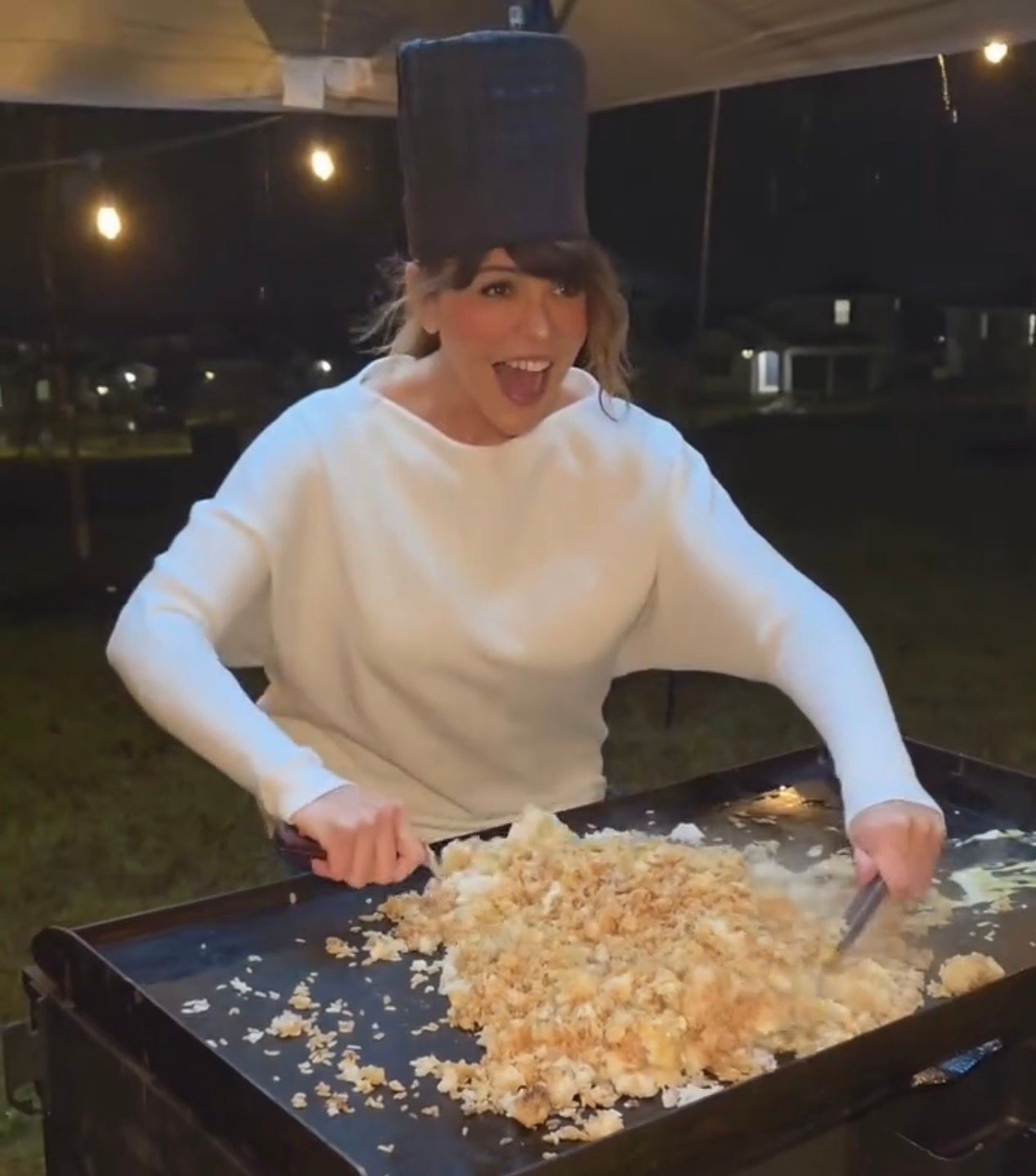 Person at a backyard nighttime cookout smiling while flipping a large pile of fried rice on a flat-top griddle under string lights, wearing a white sweater and a pot on their head.