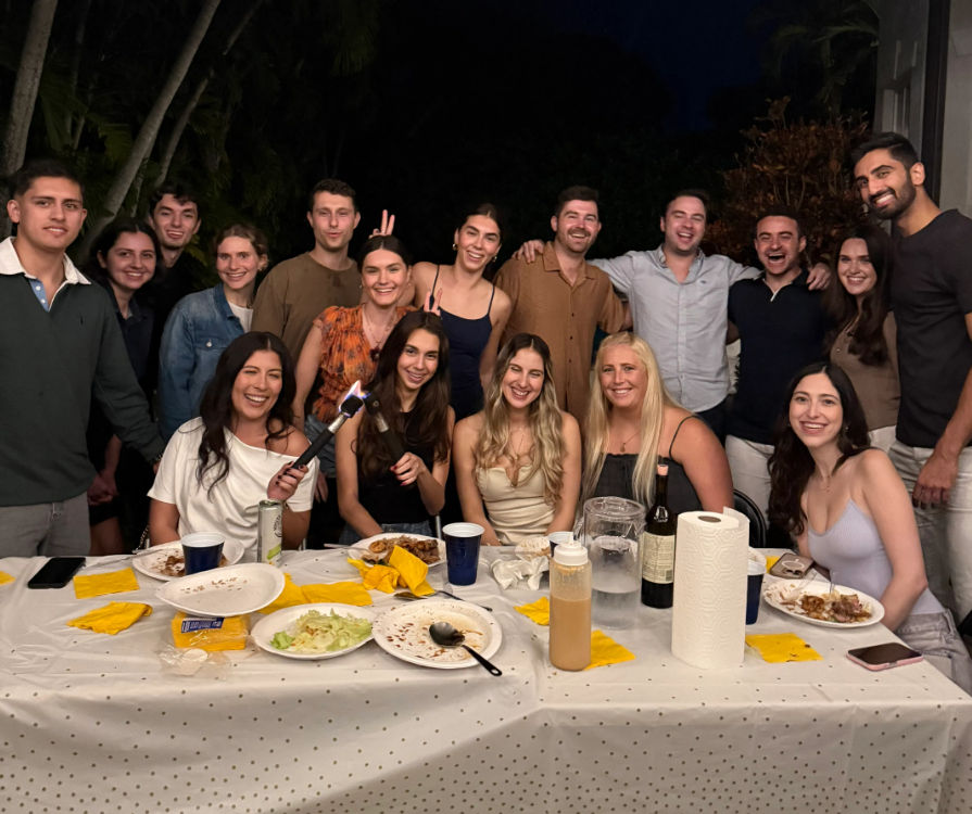 Nighttime backyard dinner party with a large group of friends smiling around a long table scattered with plates, yellow napkins, a wine bottle, paper towels and a lit handheld kitchen torch