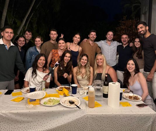 Nighttime backyard dinner party with a large group of friends smiling around a long table scattered with plates, yellow napkins, a wine bottle, paper towels and a lit handheld kitchen torch