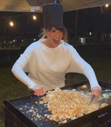 Laughing woman in a white sweater and tall hat flipping a huge mound of fried rice on a hot outdoor griddle under string lights at night