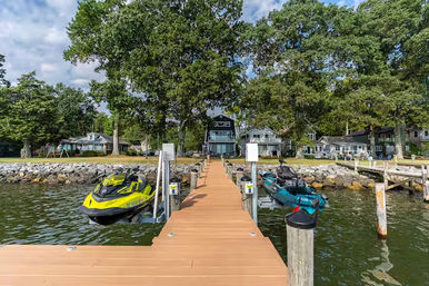 Wooden lake dock leading to lakeside homes, flanked by yellow and teal jet skis on lifts, calm green water, rocky shoreline and large oak trees on a sunny day — ready for a ride.