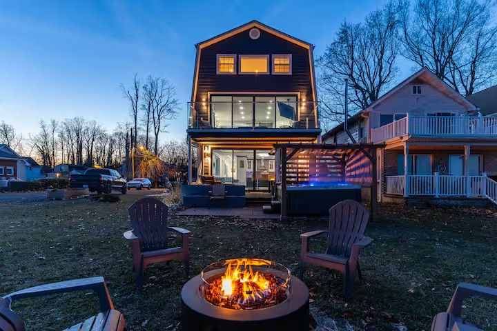 Cozy three-story modern home at dusk with illuminated glass balcony, backyard fire pit surrounded by Adirondack chairs, patio seating and hot tub.